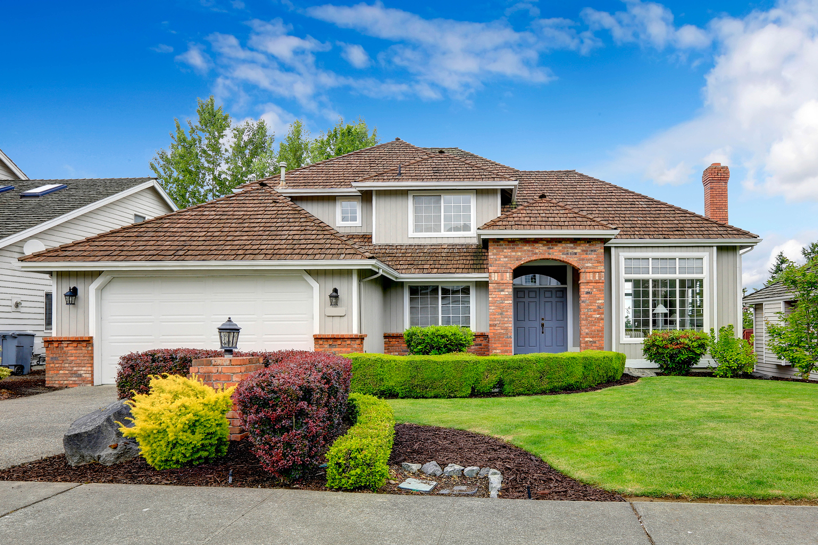 Classic house exterior with brick trimmed entrance porch green lawn and trimmed hedges. Garage with driveway